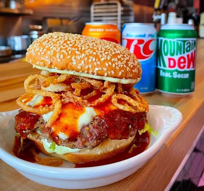 A towering burger loaded with fried onions, cheese, and sauce dripping over the patty and onto the plate below. It rests on a wooden counter with soda cans visible in the background.