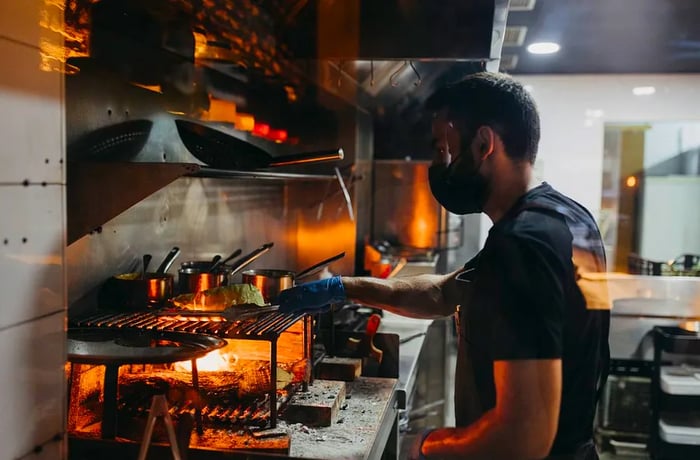 A chef wearing a black shirt and mask works at a grill station with visible flames, while wood ash is scattered across the counter.