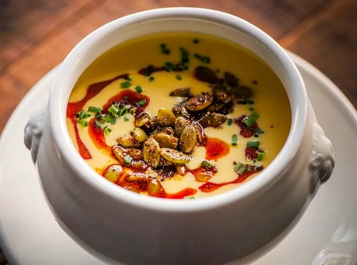 A bowl of soup served on a saucer, drizzled with sauce and topped with pepitas.