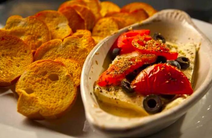 A serving of cheese garnished with tomatoes and olives, presented on a larger plate alongside toasted bread.