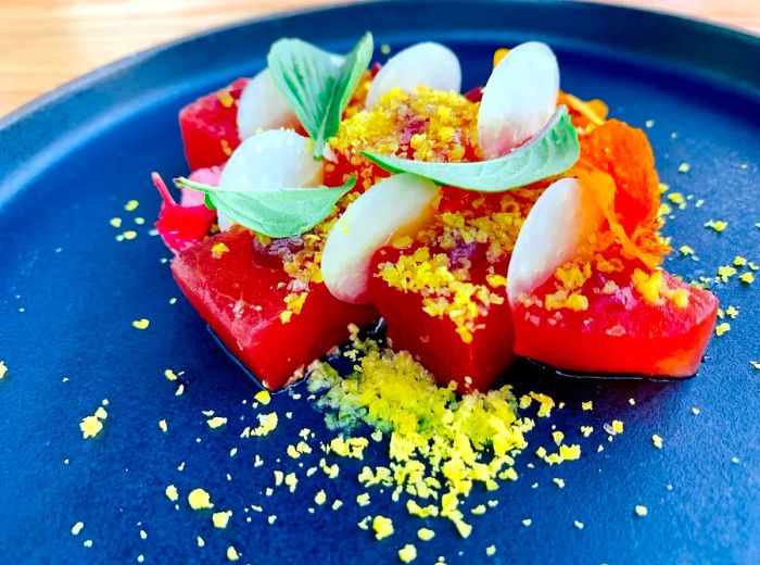 A vibrant ceramic plate holds succulent chunks of watermelon, adorned with crumbled egg yolk, slices of radish, and fresh herbs.