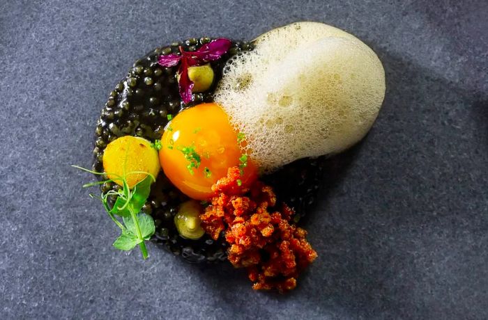 A dish featuring foam, egg yolk, caviar, and what looks like bacon bits