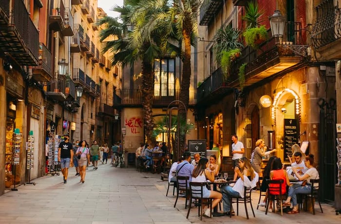 A historic cobblestone street is lined with stores selling postcards on one side, while the opposite side features outdoor tables bustling with diners.