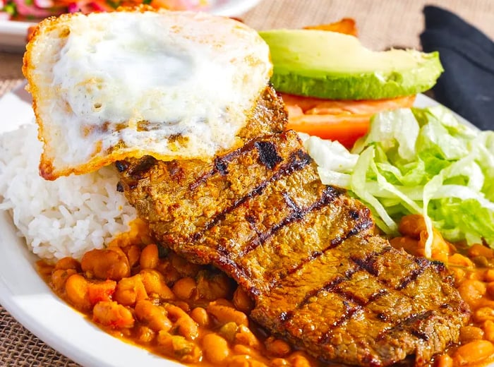 A generous grilled steak resting on a bed of beans, accompanied by fried eggs, shredded lettuce, and fresh slices of avocado and tomato.