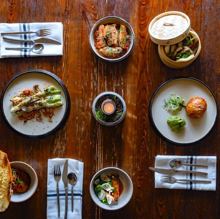 An overhead view of a wooden table adorned with place settings and a variety of dishes arranged on decorative ceramic plates and steamer boxes.