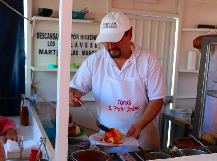 A chef in branded attire pours sauce over a torta ahogada.