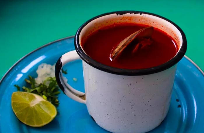 An enamel mug brimming with dark red sauce and a clam floating beside wedges of lime