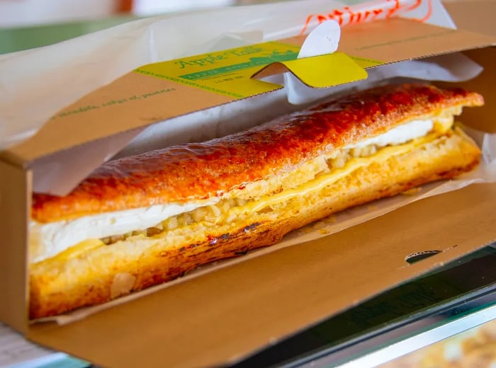 A long, sandwich-style apple strudel with its filling peeking out, presented in a branded cardboard box.