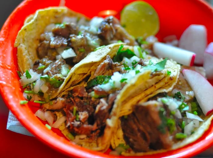 A plate of three hearty tacos, generously topped with cilantro and diced onion, accompanied by slices of radish and lime.