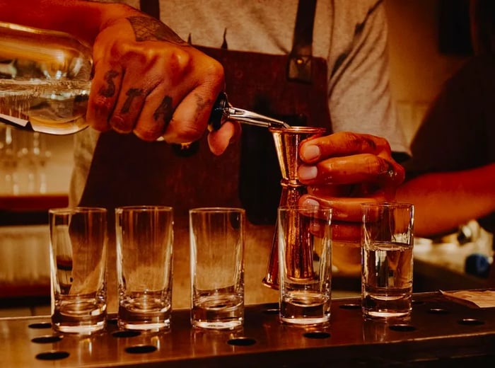 A bartender measures spirits into a jigger, preparing a lineup of drinks in the background