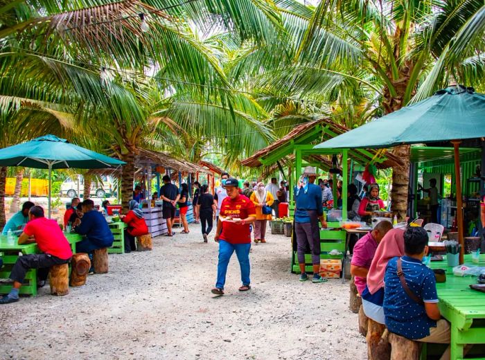 Diners enjoy meals at outdoor wooden tables, purchasing food from vendors in the background, all beneath a canopy of coconut palms.