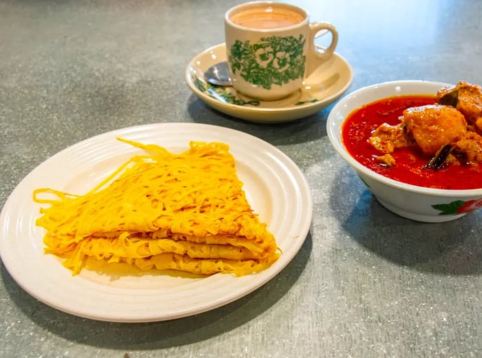 A dish of roti jala next to a cup of tea in a traditional kopitiam mug, accompanied by a stew.