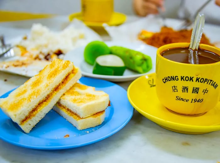 A plate stacked with halved roti bakar (coconut jam sandwich), accompanied by an assortment of desserts and a bright mug of coffee featuring the restaurant's logo.