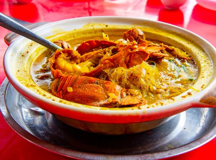 A bowl with a rim, handles, and filled with noodle soup, showcasing large prawns, resting on a saucer with a spoon protruding.