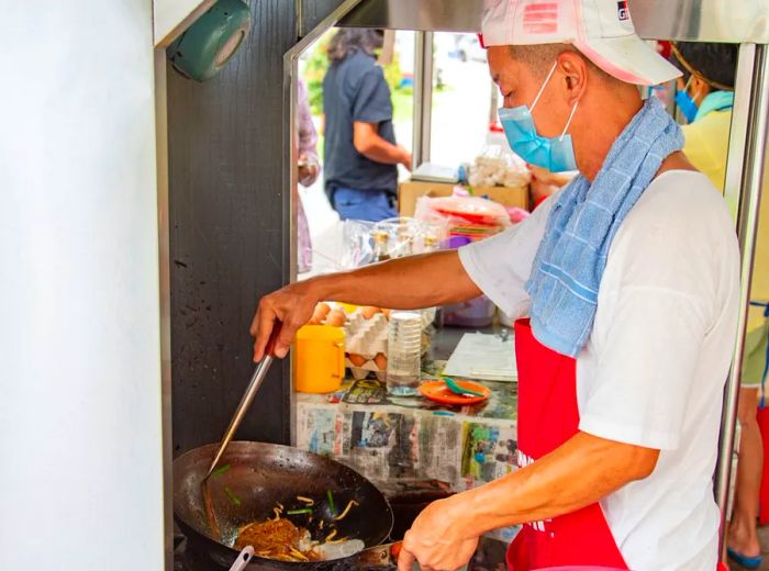 A chef wearing a surgical mask and a backward baseball cap skillfully uses a ladle in a wok.