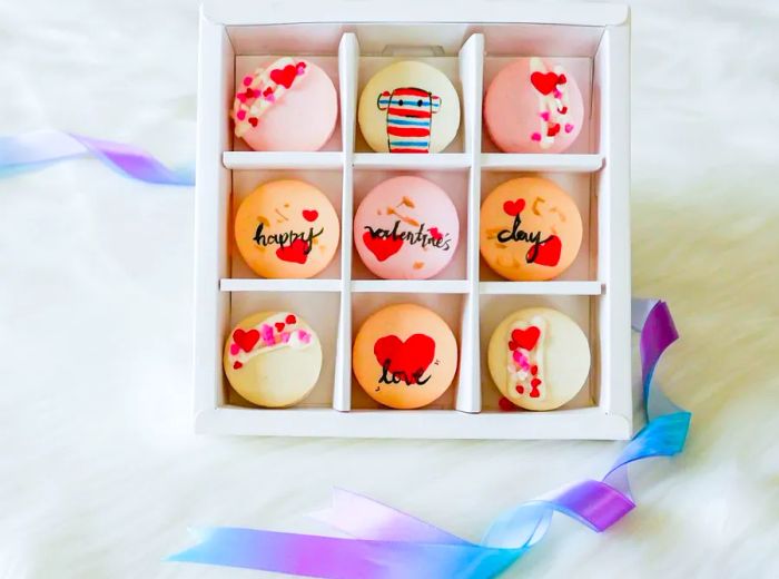 A divided box of Valentine’s Day-themed macarons in hues of white, red, and pink, adorned with heart illustrations and featuring a toy monkey.