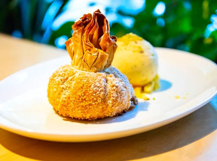 A sculptural apple strudel resembling a dumpling, adorned with a handful of paper sticking out, sits beside a small scoop of ice cream.