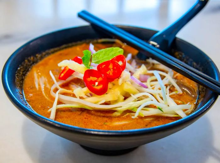 A bowl of laksa lemak adorned with chopsticks resting on the rim and a spoon at the side.