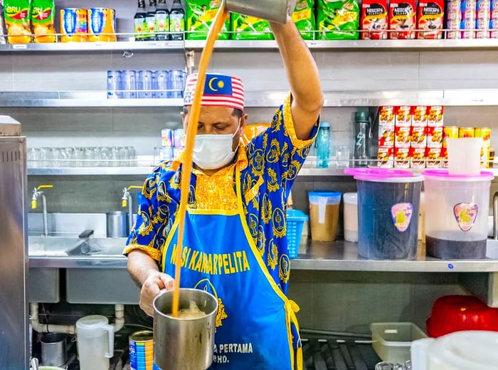 A chef, adorned in a branded apron and a cap featuring the Malaysian flag, pours tea from a metal pitcher into a pot.