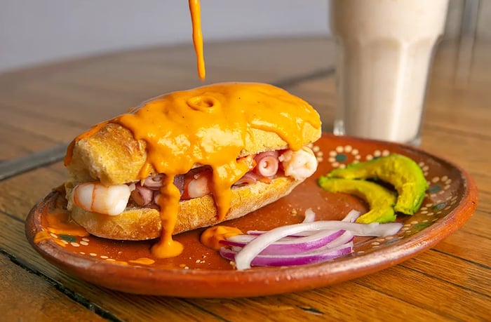 A torta filled with visible shrimp and octopus, drenched in sauce, served on a plate alongside slices of avocado and red onion.