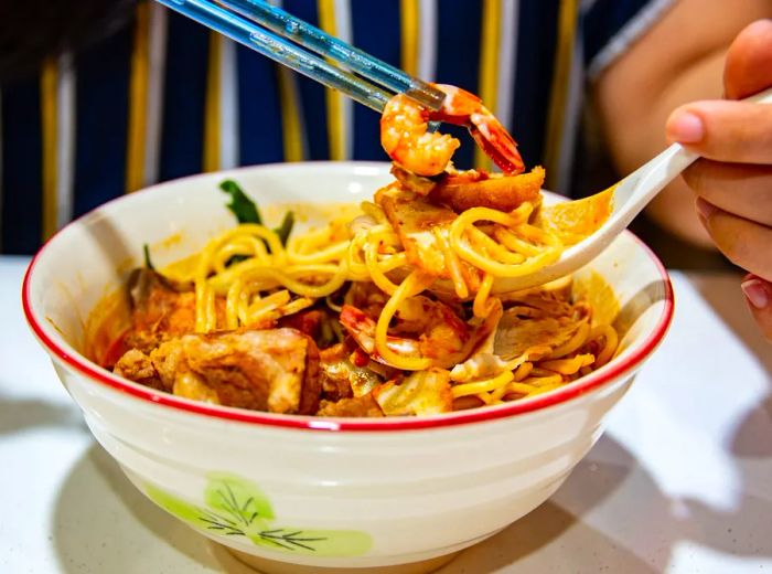 A person enjoys a bowl of har meen using both a spoon and chopsticks.