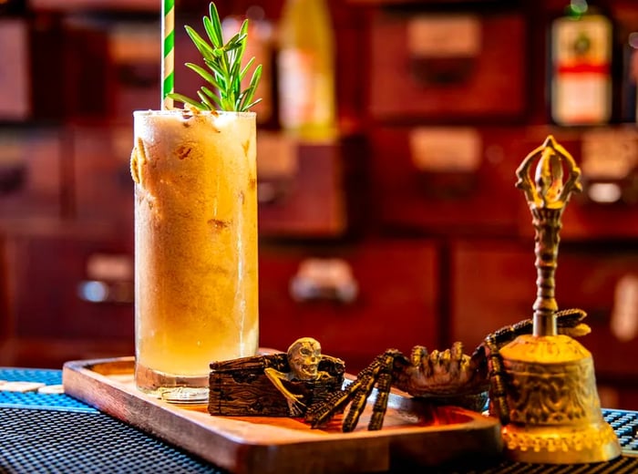 A highball cocktail adorned with herbs and actual small worms rests on a wooden platter next to a skeleton figurine inside a wooden chest, a faux spire, and an ornate bell.