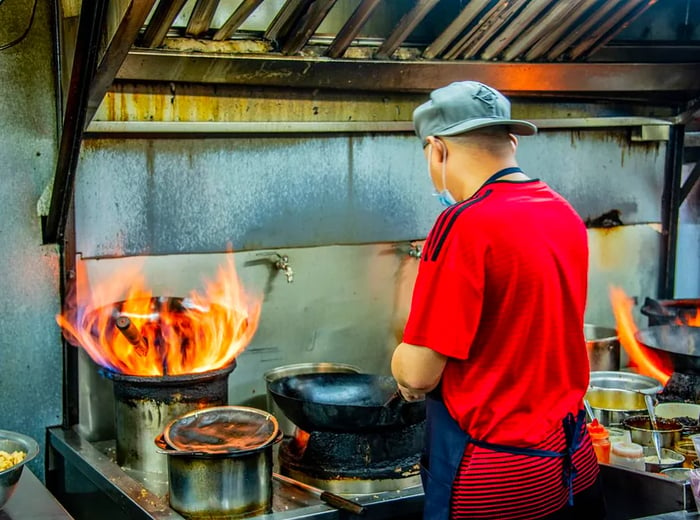 A chef dressed in a vibrant shirt and a backward baseball cap stirs a wok next to a blazing fire in another pot.