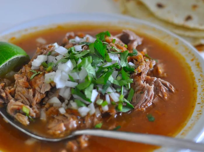 A hearty bowl of birria features a generous mound of meat rising from the rich broth, garnished with chopped onions and herbs, and a spoon poised for serving.