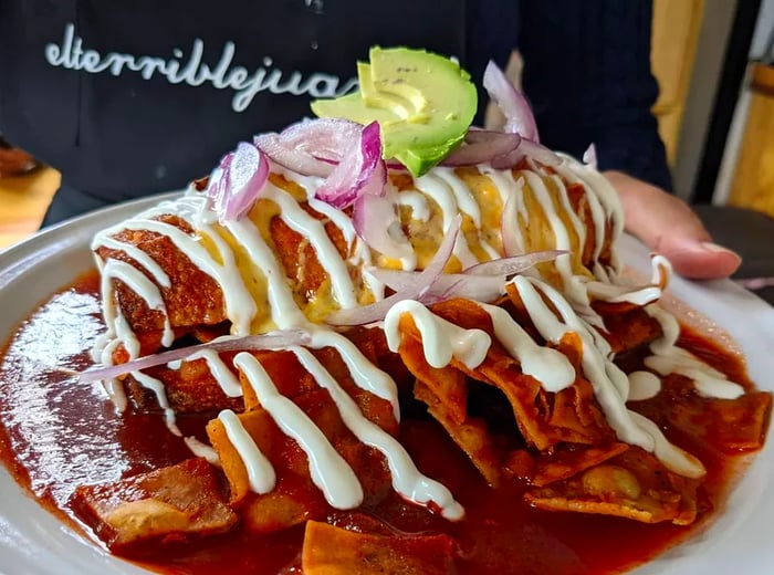 A server presents a plate overflowing with chilaquiles, drizzled with sauce and adorned with slices of avocado