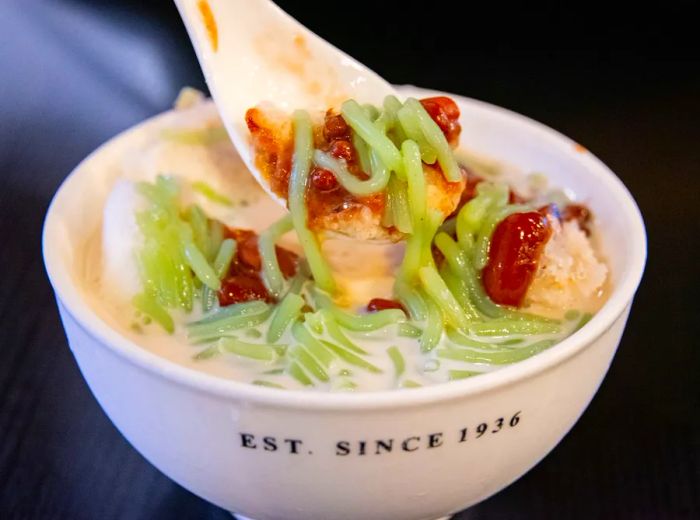 A bowl of chendol, with a spoon stirring through the noodles and toppings.