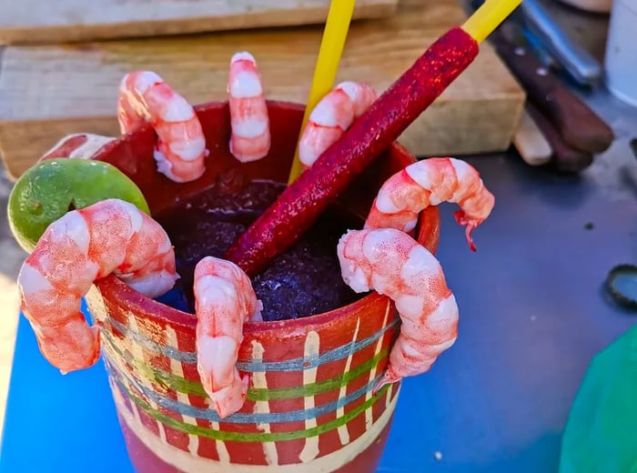 A michelada served in an artistic clay glass, adorned with fresh shrimp along the rim, a tamarind candy stick, and a wedge of lime.