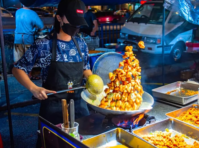 A woman wearing a Supreme hat tosses egg yolk chicken into the air from her sizzling wok.