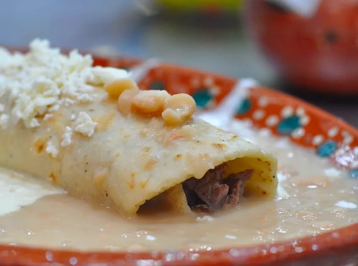 An enfrijolada stuffed with chorizo, resting in a pool of pale sauce, garnished with a few beans and a sprinkle of grated cheese