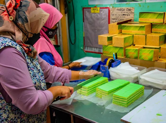 Two bakers expertly slice vibrant green pandan layer cakes using long knives.