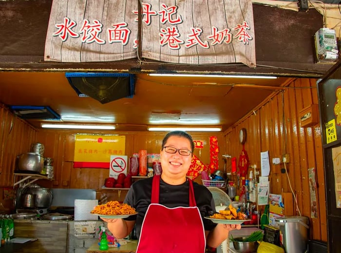 A woman stands proudly at her food stall, showcasing two delectable dishes.