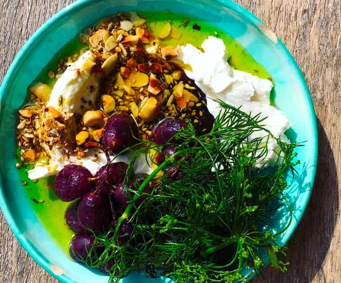 A vibrant ceramic plate showcasing a generous mound of labneh garnished with dukkah, accompanied by dark grapes and fresh greens.