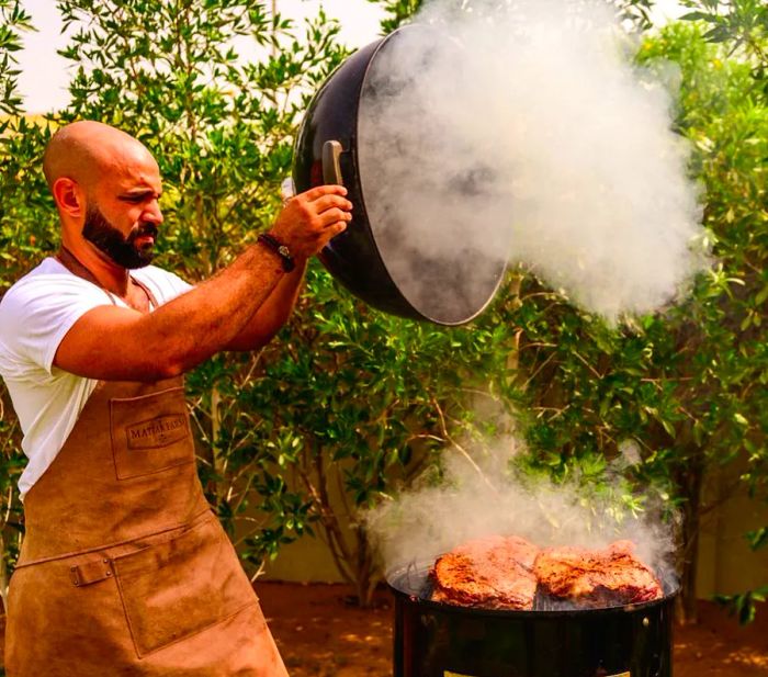 A pitmaster lifts the grill lid, releasing a cloud of smoke over large cuts of meat as they cook.