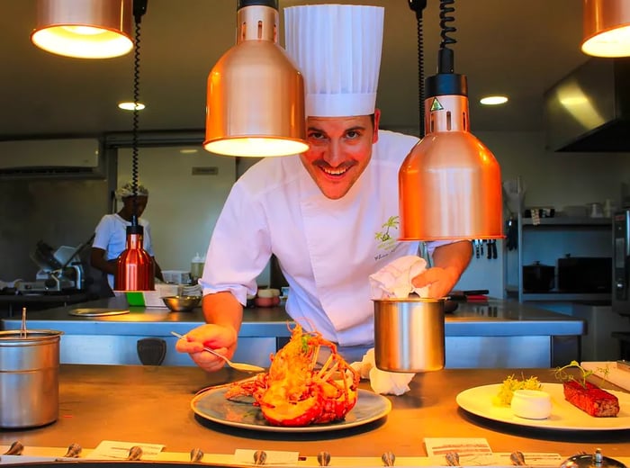 A chef dressed in whites and a toque examines a dish featuring spiny lobster.