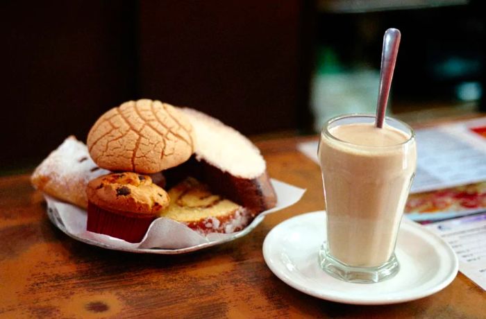 A plate of assorted simple pastries sits alongside a tall glass of creamy coffee and a spoon.