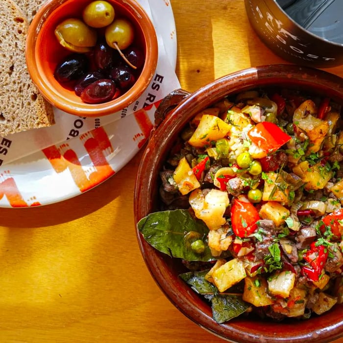 A vibrant grain salad featuring chunks of potato, bell pepper, peas, and fresh herbs, elegantly served in an earthenware bowl alongside a smaller bowl of olives.