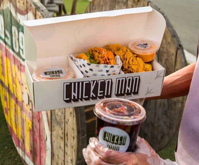 A person wearing kitchen gloves holds a takeout box filled with fried chicken and a drink in a to-go cup