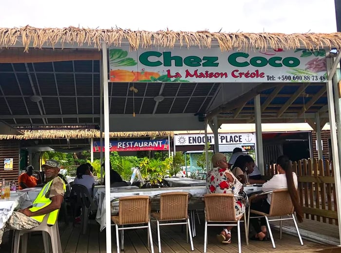 A vibrant open-air restaurant beneath a palm frond roof features a prominent banner displaying the name Chez Coco, with diners enjoying their meals at tables.