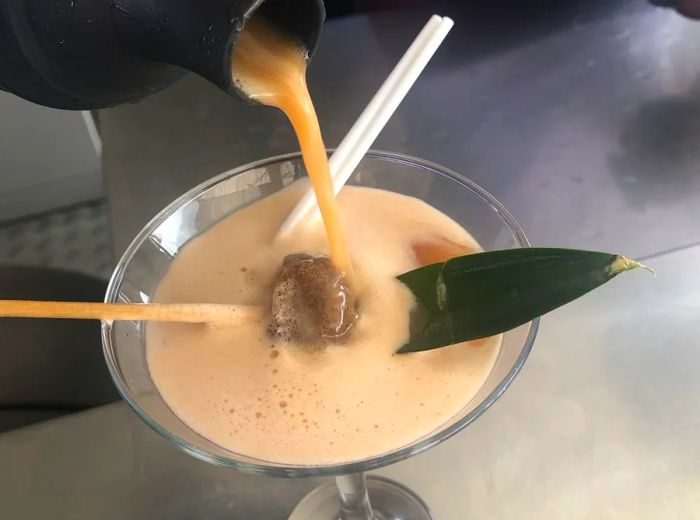 A bartender pours a rich cocktail from a shaker into a martini glass, adorned with a piece of tamarind candy on a skewer and a tropical leaf.