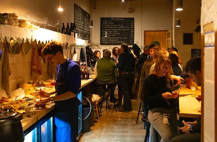 Diners enjoy their meals at elevated tables along one wall, with a long restaurant layout featuring a chef preparing dishes to one side and large chalkboard menus displayed on the opposite wall.