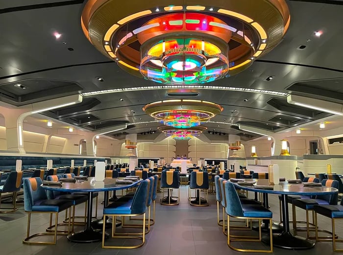 An expansive, futuristic dining hall featuring blue chairs and an eye-catching colorful chandelier.