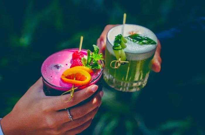 Cheers erupt as people clink vibrant cocktails—one pink adorned with a skewer of berries and lemon, the other green, topped with fresh herbs