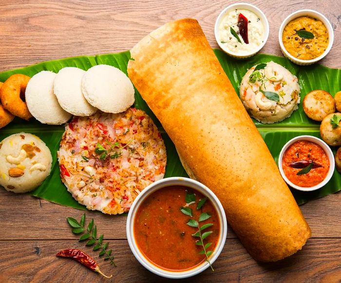 From above, an assortment of dishes arranged around a large dosa, presented on a leaf atop a wooden table