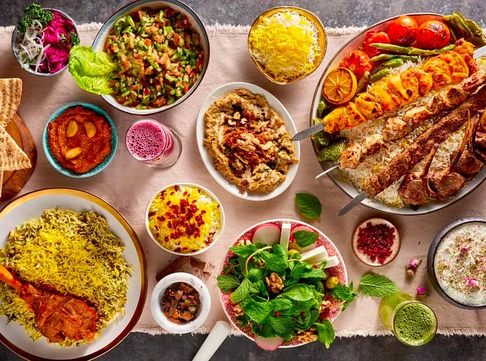 A long table filled with an array of dishes, including a large platter of kebabs served over saffron rice, a leg of lamb, fresh salad, and flatbread with dips.