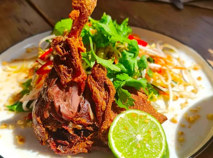 A slightly shredded duck leg, displayed upright on a plate, accompanied by a vibrant herb salad and a lime wedge.