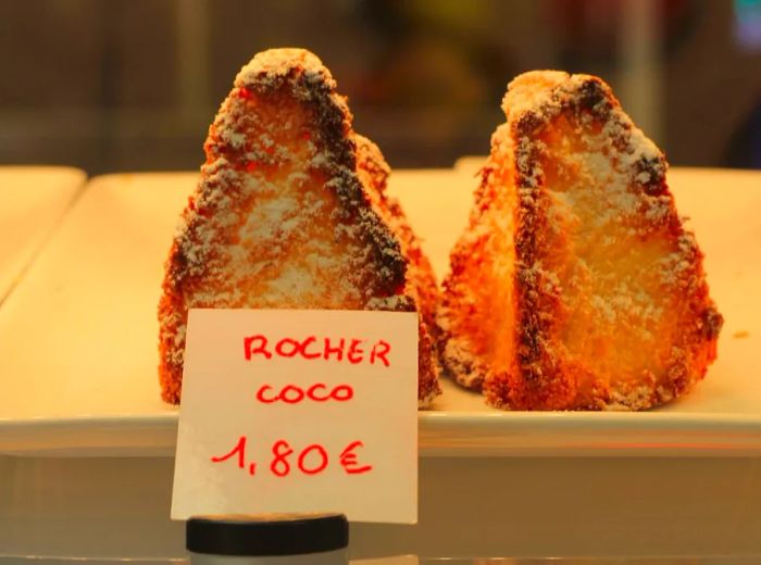 Sugar-dusted macaroons displayed in a pastry case with a handwritten sign reading ‘rocher coco’